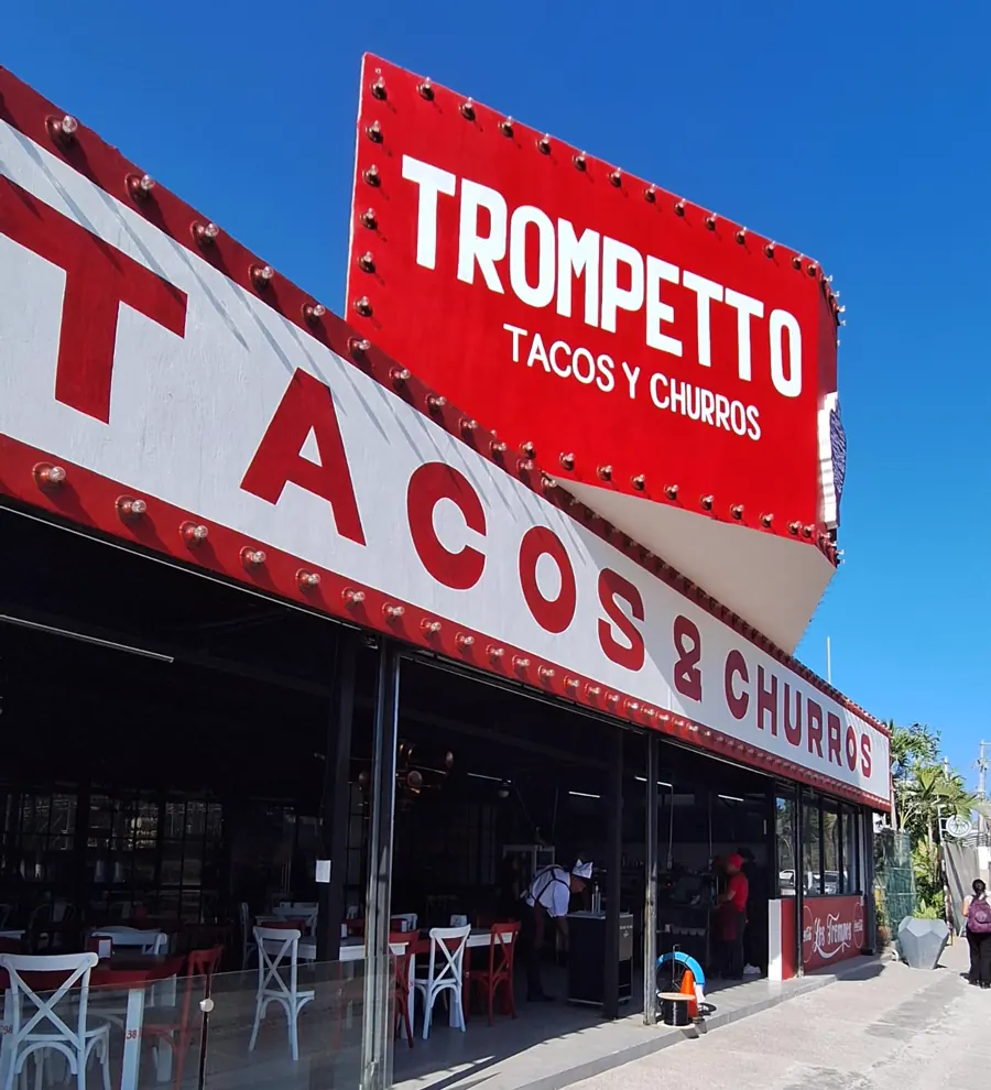 Fachada de Trompetto: letrero rojo con focos que dice Trompetto, tacos y churros, y el toldo con las mesas rojas y blancas debajo