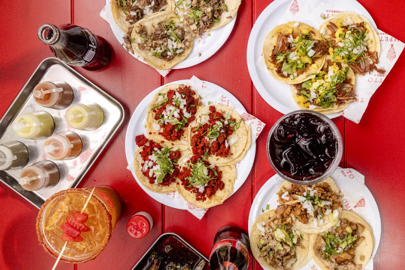 Vista desde arriba de una mesa roja de Trompetto con varios platos de tacos, salsas en botella y bebidas