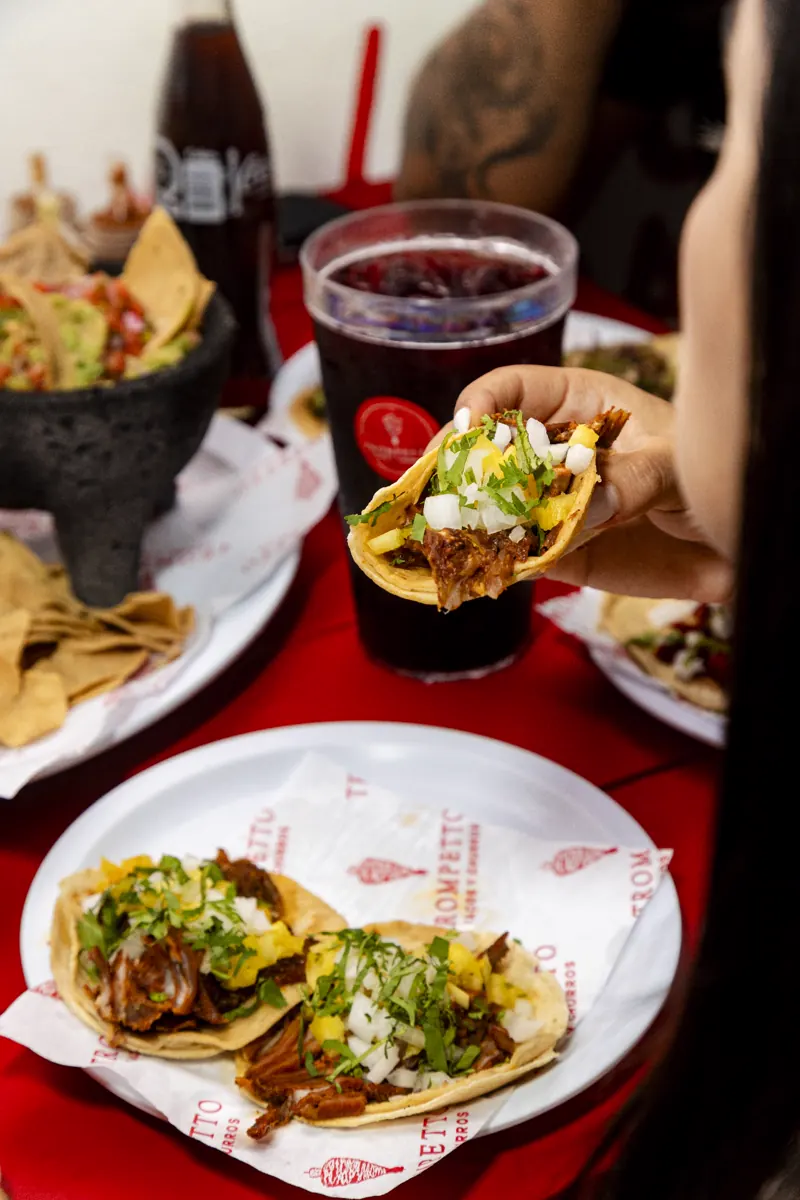 Una mano toma un taco con cebolla y cilantro sobre una mesa roja, junto a un molcajete con guacamole y un vaso de agua de jamaica
